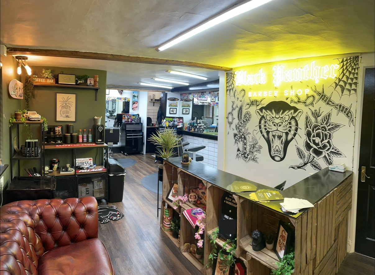 Inside Black Panther Barbershop: the yellow neon Black Panther Barber Shop sign over a black and white panther mural, a chesterfield sofa, the coffee bar shelf and the barber chairs behind